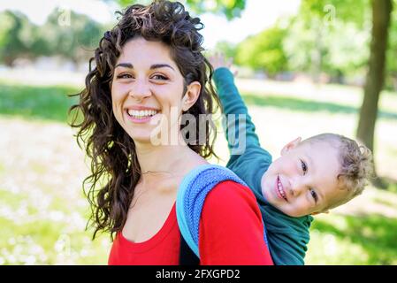 Nahaufnahme Porträt einer glücklichen jungen Mutter trägt ein Kind auf dem Rücken, gebunden mit einem bunten traditionellen Stück Stoff in einem öffentlichen Park Garten. Mama Stockfoto