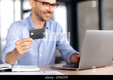 Online-Shopping-Konzept. Verschwommener junger glücklicher Kaukasusmann, der eine Brille trägt, in stilvoller Kleidung sitzt, am Tisch sitzt, Laptop und Kreditkarte verwendet, um für Online-Einkäufe zu bezahlen oder Waren nach Hause zu liefern Stockfoto