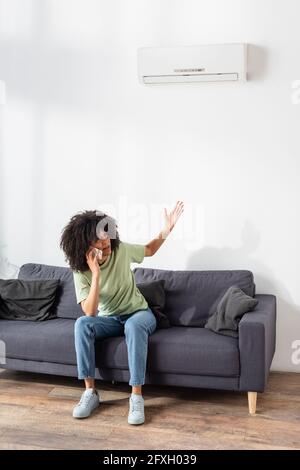 Unzufrieden afroamerikanische Frau, die auf dem Smartphone spricht und darauf zeigt Kaputte Klimaanlage im Wohnzimmer Stockfoto
