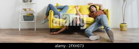 Erschöpfte junge multiethnische Paare, die auf dem gelben Sofa liegen und im Sommer unter Hitze leiden, Banner Stockfoto