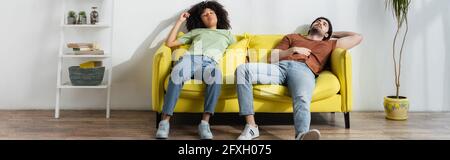 Erschöpfte multiethnische Paare sitzen auf dem gelben Sofa und leiden im Sommer unter Hitze, Banner Stockfoto