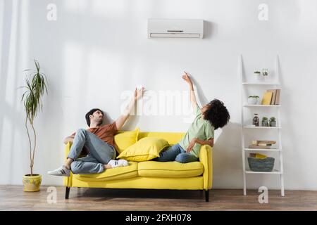 Multiethnisches Paar, das auf dem gelben Sofa sitzt und die Klimaanlage erreicht Während sie im Sommer unter Hitze leiden Stockfoto