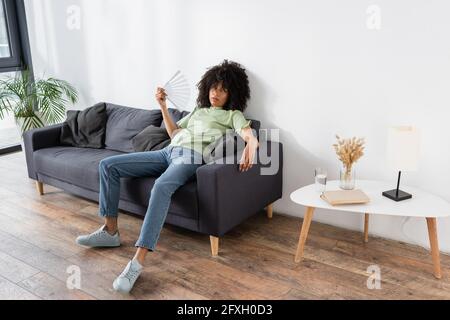 afroamerikanische Frau winkt mit Handventilator, während sie auf dem Tisch sitzt Graue Couch und leiden unter Hitze Stockfoto