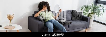 afroamerikanische Frau winkt mit Handventilator, während sie Wasser trinkt und auf der grauen Couch sitzt, Banner Stockfoto