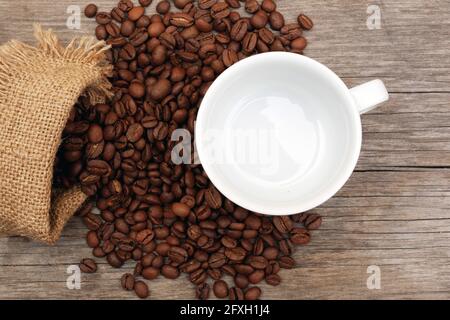 Draufsicht auf Kaffeebohnen, die aus Sackleinen verschüttet wurden und Weiße leere Tasse auf veraltertem Holzhintergrund Stockfoto
