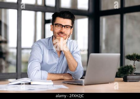 Porträt ein hübscher junger kaukasischer Freiberufler oder Manager, der striktes Hemd und Brille trägt, mit einem Laptop am Tisch sitzt, seinen Kopf mit der Hand stützt, die Kamera anschaut und freundlich lächelt Stockfoto