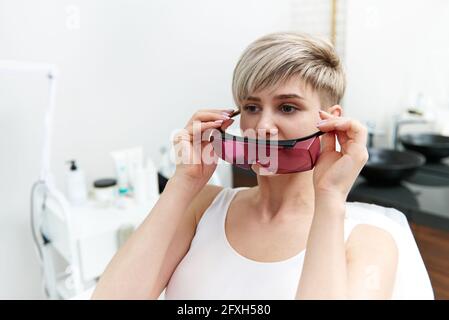 Attraktive blonde Frau sitzt auf einem medizinischen Stuhl im Kosmetologie-Zimmer Im Spa-Salon und das Anziehen einer UV-Schutzbrille Stockfoto