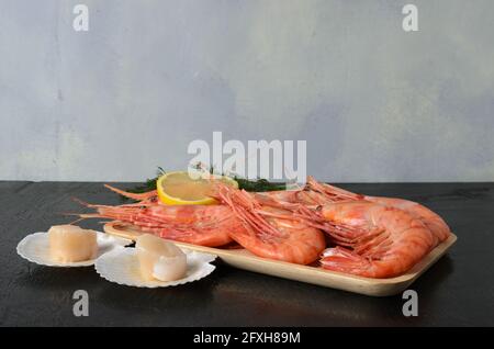 Gekochte Garnelen in einer Schale mit Dill und Zitrone in einer Holzschüssel mit einer rohen Jakobsmuschel in einer Schale auf dunklem Holzgrund. Selektiver Fokus. Stockfoto