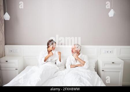 Verärgeres Ehepaar in weißen Bademänteln, die zueinander schauen Beim Gestik in der Wohnung Stockfoto