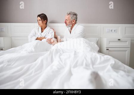 Traurige gestresste Frau im weißen Bademantel schaut weg, während sie sitzt Auf dem Bett in der Wohnung Stockfoto