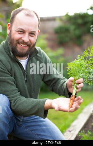 Portrait glücklicher Mann mit Bart zeigt frisch geerntete Karotte im Garten Stockfoto