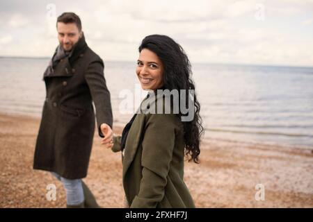 Portrait glückliches Paar in Wintermänteln, das die Hände am Strand hält Stockfoto