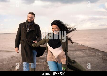 Glückliches Paar in Wintermänteln, das die Hände am Meeressteg hält Stockfoto