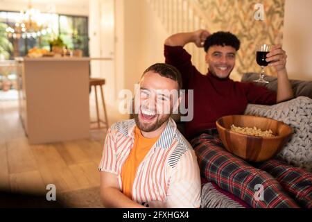 Glückliches schwules männliches Paar mit Wein und Popcorn, lachend und fernsehend Stockfoto