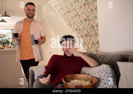 Glückliches schwules männliches Paar mit Rotwein und Popcorn, das zu Hause Fernsehen schaut Stockfoto