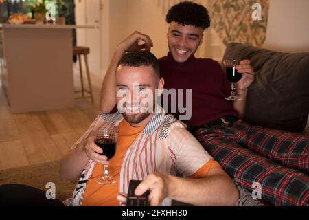 Glückliches schwules männliches Paar, das Wein trinkt und auf dem Sofa Fernsehen schaut Stockfoto