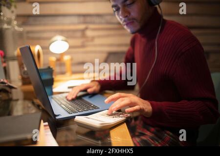 Junger Mann mit Kopfhörern, der im Heimbüro einen Rechner am Laptop benutzt Stockfoto
