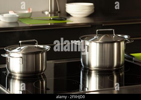 2 Töpfe mit Deckel befinden sich auf einem Herd in der Küche und spiegeln sich in ihnen wider. Im Hintergrund befinden sich kleine Teller, Schüsseln und ein Messer. Stockfoto