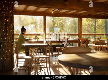 Geschäftsfrau mit Kopfhörern, die in einem sonnigen Restaurant am Laptop arbeitet Stockfoto