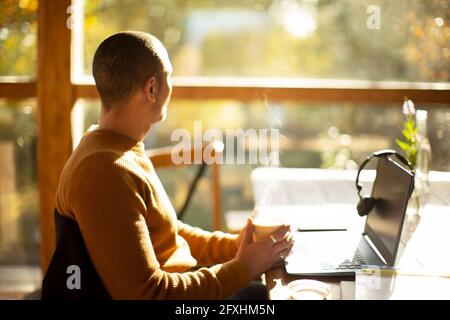 Durchdachter Geschäftsmann mit Kaffee und Laptop und Blick aus dem sonnigen Fenster Stockfoto