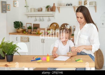 Mutter und Tochter machen Hausaufgaben. Ein kleines Mädchen absolviert nachdenklich Matheaufgaben unter der Aufsicht eines Tutors. Das Konzept der Auto nehmen Stockfoto