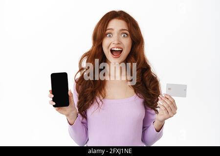 Aufgeregt Mädchen mit roten lockigen Haaren, keuchend verwundert und erstaunt, zeigt Smartphone leeren Bildschirm und Kreditkarte, demonstriert Online-Shop oder Bank Stockfoto