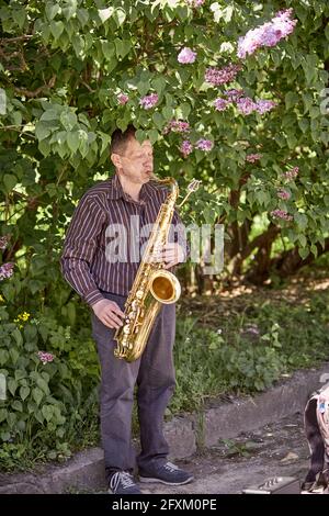 Kiew, Ukraine - 16. Mai: Ein Straßensaxophonist spielt an einem sonnigen Tag in einem Frühlingspark Trompete. Tag frei, Unterhaltung für die Menschen. Vertikales Foto Stockfoto