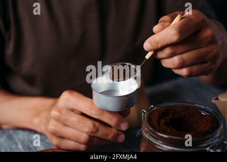 Ein junger Kaukasier füllt den Trichter einer Mokakanne mit gemahlenem Kaffee und sitzt an einem grauen rustikalen Holztisch Stockfoto