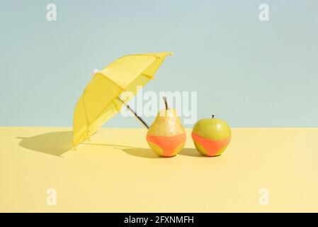 Sommerliches Fruchtkonzept. Nasser Apfel und eine Birne in Monokini neben einem Regenschirm isoliert auf einem blau-gelben Hintergrund. Zusammenfassung. Rechtecklayout mit c Stockfoto