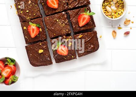 Schokoladenquadrate mit Pistazien und Erdbeeren auf weißem Papier auf hellem Hintergrund, Draufsicht, horizontale Komposition. Flache Lebensmittel Stockfoto