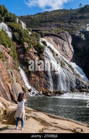 Kaukasische Frau fotografiert den Ezaro-Wasserfall mit Wasser, das zwischen Felsen auf dem See in Galicien, Spanien, Europa zusammenbricht Stockfoto
