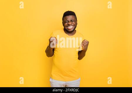 Kopf geschossen Studio-Portrait euforisch lachend glücklich afroamerikanischen Mann in gelben T-Shirt klammernden Fäusten, machen ja Geste, den Sieg zu feiern Stockfoto