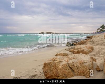 Leerer malerischer Nissi-Strand mit aufgewühlten türkisfarbenen Wasser, einer kleinen felsigen Insel und einem dunklen, wolkigen Himmel in Ayia Napa, Zypern. Herrlicher Urlaub am Meer. Stockfoto