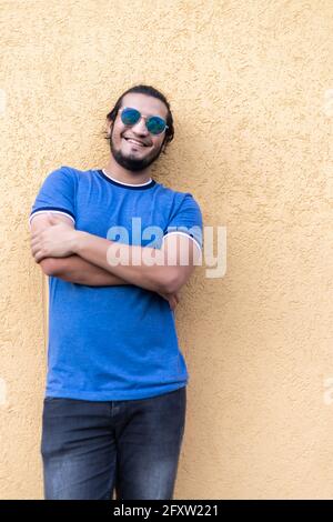 Der junge Latino-Mann trägt Sonnenbrille und lässige Kleidung, die an der gelben Wand steht und selbstbewusst mit gekreuzten Armen lächelt. Frische Luft Stockfoto