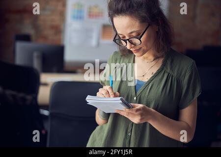 Kreative Innenarchitektin oder Architektin in einer Brille, die Notizen in ihrem Notizbuch macht, über ein neues Projekt nachdenkt, in ihrem Büro auf einem steht Stockfoto