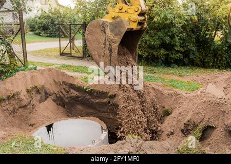 Ein Eimer Bagger mit einem Haufen Sand und Erde begräbt Kanalisation Betonringe in der Industriezone. Stockfoto