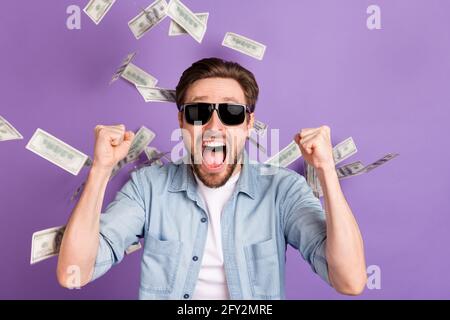 Foto von beeindruckt glücklich jungen Kerl gekleidet Denim Hemd dunkel Brille Geld fliegen steigende Fäuste isoliert lila Farbe Hintergrund Stockfoto