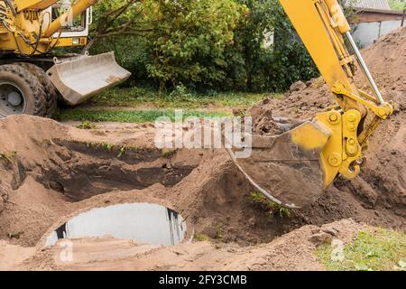 Ein Eimer Bagger mit einem Haufen Sand und Erde begräbt Kanalisation Betonringe in der Industriezone. Stockfoto