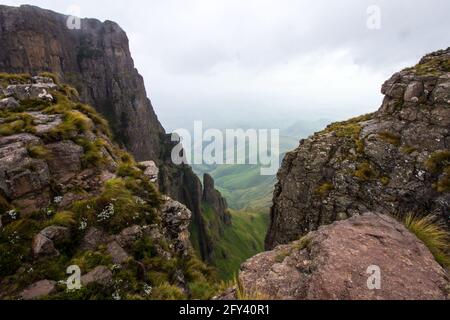 Blick über den Rand auf Castle Buttress, in den Drakensberg Mountains in Südafrika, mit dem Nebel beginnt sich zu heben, um die grünen Hügel unten zu offenbaren. Stockfoto