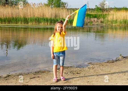 Vor der Kulisse des Sees flattert die Flagge der Ukraine in den Händen eines kleinen Mädchens. Unabhängigkeitstag der Ukraine und Flaggentag. Stockfoto