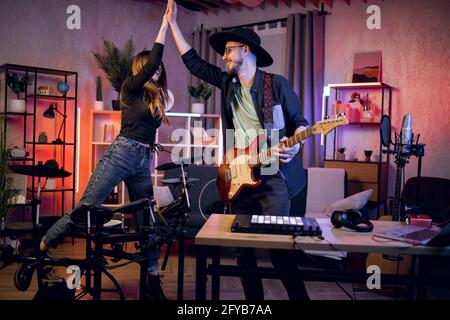 Gutaussehender Mann und charmante Frau, die sich nach einer erfolgreichen Probe im Heimstudio hohe fünf gaben. Zwei glückliche Musiker, die im Innenbereich elektronische Gitarre und Schlagzeug benutzen. Stockfoto