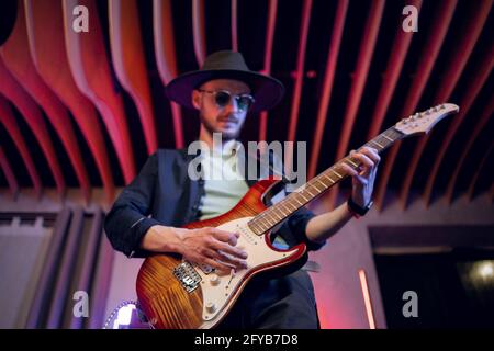 Blick aus der Perspektive eines talentierten Musikers, der trendige Hüte und Sonnenbrillen trägt und in Innenräumen E-Gitarre spielt. Konzept von Menschen, Musik und kreatives Hobby. Stockfoto