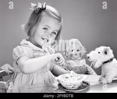 1940ER-JAHRE LÄCHELNDES MÄDCHEN BOGEN IM HAAR ESSEN SCHÜSSEL EIS BEIM BLICK AUF DIE KAMERA, UMGEBEN VON AUSGESTOPFTEM TIERHUND UND PUPPENSPIELZEUG - F2693 HAR001 HARS JOY LIFESTYLE WEIBCHEN GEWINNER STUDIO SHOT STOPFTE GESUNDHEIT ZU HAUSE LEBEN KOPIEREN RAUM HALBE LÄNGE B&W HUMORVOLLES GLÜCK FRÖHLICH UND ERNÄHRUNG, KOMISCH VON EINEM LÄCHELN UMGEBEN COMEDY VERBRAUCHEN FREUDIGE NAHRUNG STILVOLLE EIS ANGENEHM ANGENEHM ANGENEHM CHARMING GROWTH JUVENILES LIEBENSWERT LIEBENSWERT BEZAUBERND ANSPRECHEND SCHWARZ UND WEISS KAUKASISCHE ETHNIZITÄT HAR001 ALTMODISCH Stockfoto