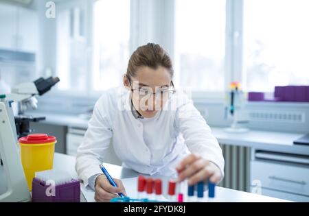 Frau im Labor experimentiert mit dem Halten der Ampulle. Junger Wissenschaftler, der sich Notizen über die Ergebnisse von Forschungstests gemacht hat. Stockfoto