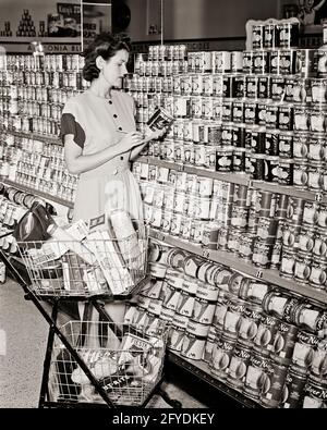 1940ER BRÜNETTE FRAU MIT EINKAUFSWAGEN IM LEBENSMITTELGESCHÄFT GANG LESEN DES ETIKETTS AUF DER DOSE FRUCHTSAFT - S11549 HAR001 HARS BRUNETTE HAUSFRAU KÄUFER LABEL HAUSFRAUEN WAREN WAHL PRÜFUNG HAUSFRAUEN GESCHÄFTE ENTSCHEIDUNG INSPEKTION STILVOLLE COMMERCE JUNGE ERWACHSENE FRAU SCHWARZ UND WEISS UNTERNEHMEN KAUKASISCHE ETHNIZITÄT HAR001 SAFT ALTMODISCH AUSWAHL Stockfoto