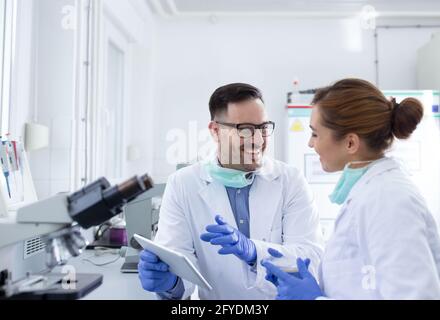 Mann und Frau Labortechniker Ärzte halten Tablet und experimentieren mit dem Mikroskop. Zwei junge Wissenschaftler diskutieren Ergebnisse und experimentieren im Labor Stockfoto