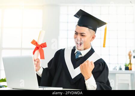 Glückliche männliche Abschluss beobachten den Laptop und hält Diplom und Erfolg wird gefeiert Stockfoto