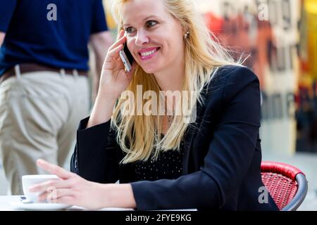 Reife Frau, die Telefon in City Street Cafe vor Aus dem Schaufenster, um eine Tasse Kaffee zu trinken Stockfoto