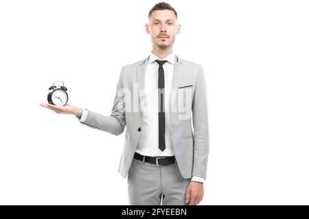 Horizontales mittleres Porträt eines hübschen jungen erwachsenen kaukasischen Mannes mit grauem Anzug mit Wecker, Deadline-Konzept, weißem Hintergrund Stockfoto