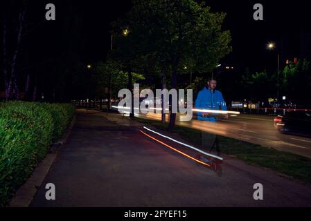 Kaukasischer Mann, der in der Nacht auf der Gasse einen Elektroroller fährt. Verschwommenes Licht und lange Belichtung Stockfoto